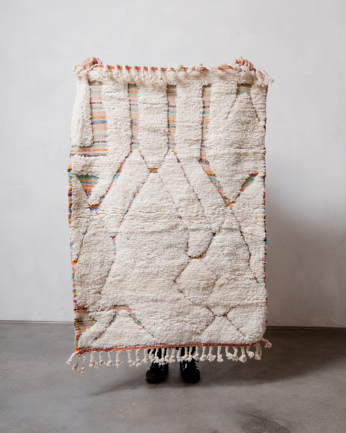 Small Berberlin Beniourain rug with cream wool pile and woven base of multicoloured stripes, being held up in front of a white wall.